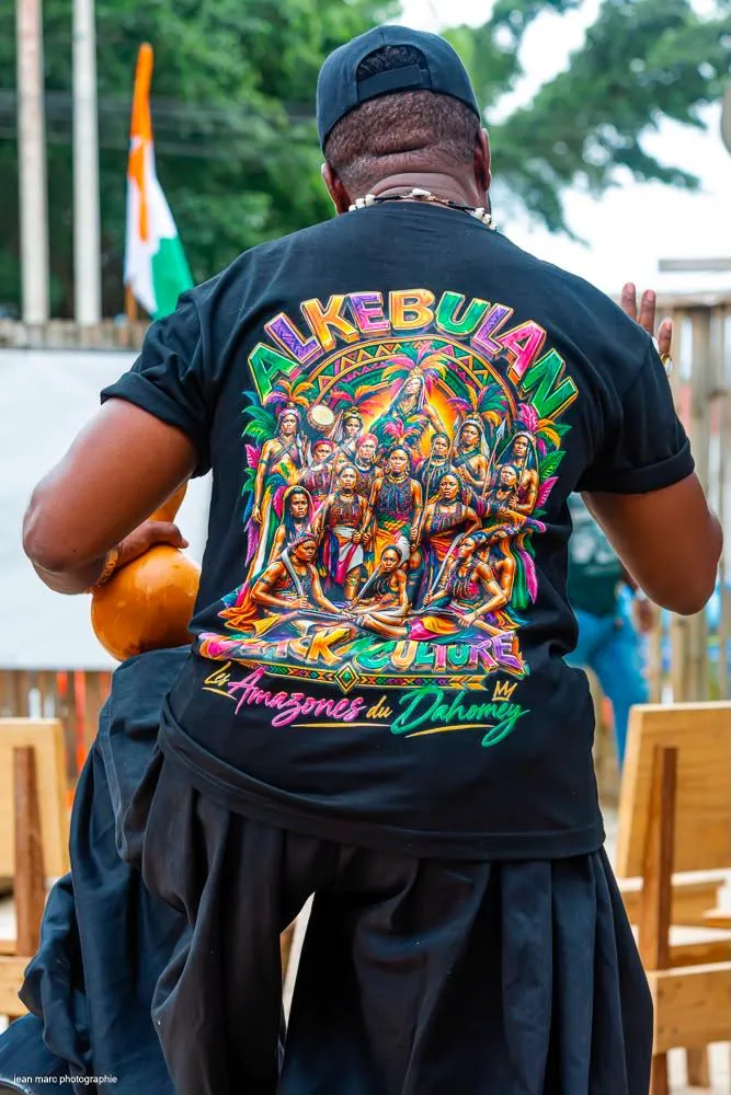 Homme de dos portant le t-shirt Amazones du Dahomey, drapeau ivoirien en arrière-plan