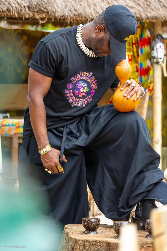 Homme en t-shirt logo soleil multicolore et sarouel noir, une calebasse à la main